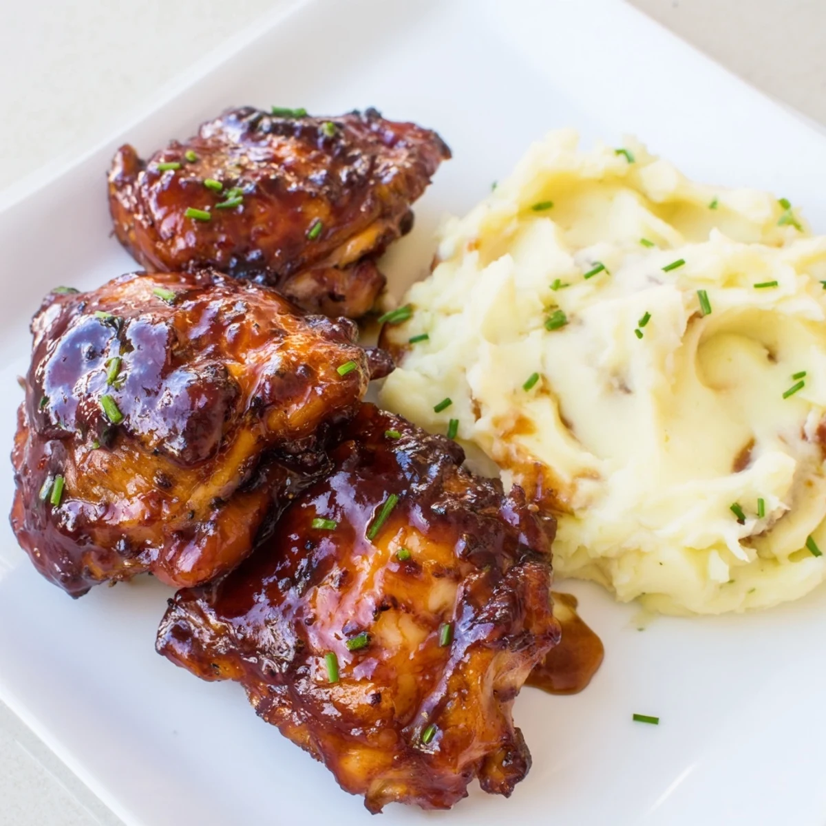 Close-up of baked honey BBQ chicken thighs, the sauce bubbling, on a plate with fluffy mashed potatoes.